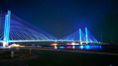 A brightly lit suspension bridge at night, illuminated with blue lights, reflecting on the water below.